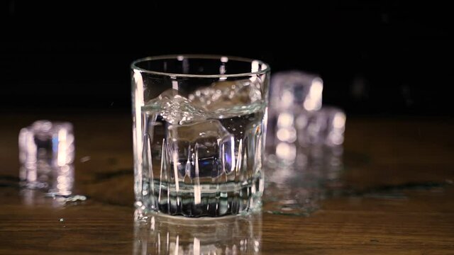 Two ice cubes drop into glass of water slashing out onto wood counter in slow motion