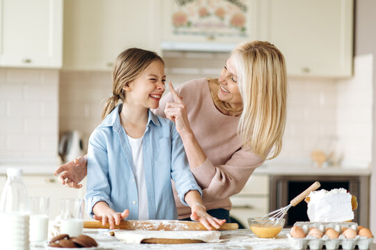 Cheerful mom or grandma and her pretty little daughter or granddaughter spend time together at the kitchen. They cook together a pie, indulge in flour, laugh and have fun