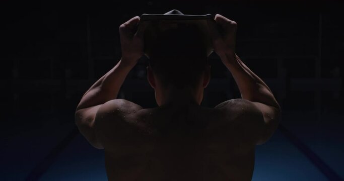 Cinematic back view shot of young professional male swimmer is putting on swim cap before to start practicing and training with effort in swimming pool. Concept of sport, competition, victory, fitness