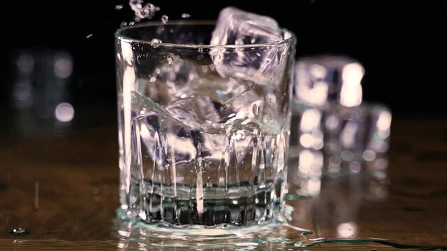 Slow pan in of ice cubes dropping into glass of water splashing onto wood counter in slow motion
