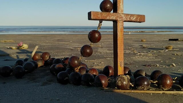 A cross wrapped in rosary beads sits on a beautiful beach with waves in the background.