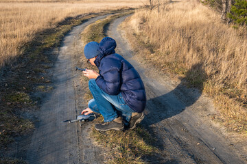 A male tourist with a remote control sets up a drone on a dirt road in a field on a clear sunny autumn day.