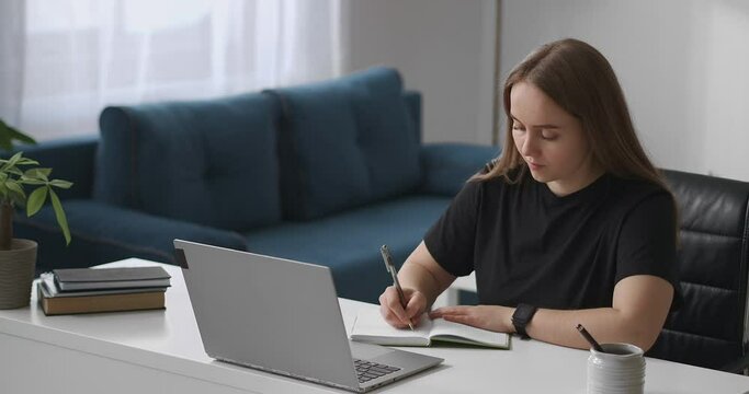 young student woman is listening to lecture by video in laptop and writing notes in notebook, distant education and e-learning, medium portrait