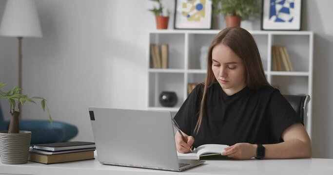 young woman is reading information on screen of laptop and writing in notebook, learning in home, distant education, online course