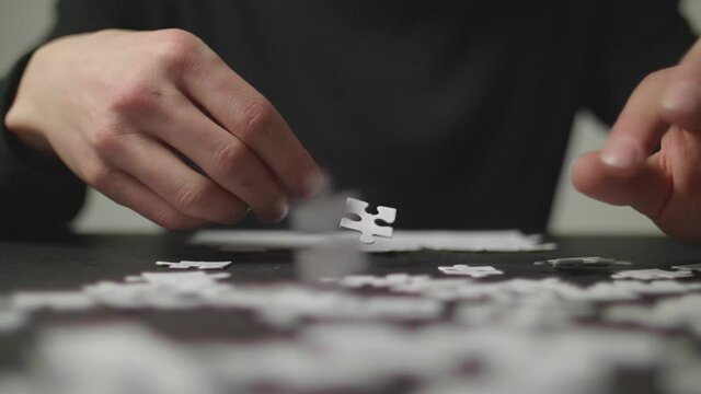 Person's hands trying to solve a puzzle in the background with white puzzle pices falling on a dark table simultaneously. Shot in 4K, Slow Motion with Shallow Depth of Field.	
