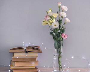 Eustoma flowers bouquet in a vase, stack of old vintage books and garland lights on a grey background. Reading and relax concept.