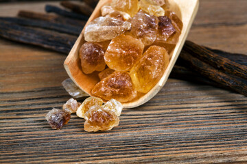 Crystals of brown sugar in a wooden scoop with vanilla beans on a wooden background.