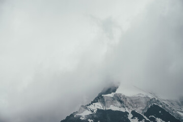 Fototapeta na wymiar Minimalist monochrome atmospheric mountains landscape with big snowy mountain top in low clouds. Awesome minimal scenery with glacier on rocks. Black white high mountain pinnacle with snow in clouds.