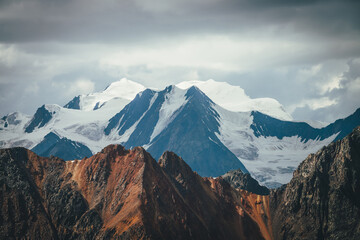Atmospheric mountains landscape with great snowy top behind colorful brown red orange rocky wall under cloudy sky. Dramatic highland scenery with giant glacier and big vivid brown red orange mountain.