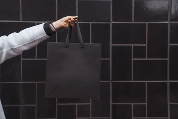 Woman holds in hand black shopping bag against black textured tile wall. Blank space, minimal brand...