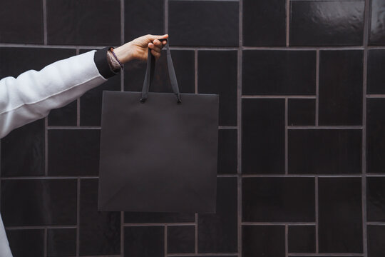 Woman holds in hand black shopping bag against black textured tile wall. Blank space, minimal brand package mock up concept, fashion blogger, space for text