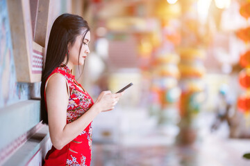 Portrait beautiful smiles Asian young woman wearing red traditional Chinese cheongsam decoration and writing message on smartphone for Chinese New Year Festival at Chinese shrine in Thailand