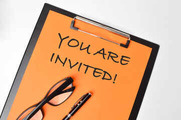 Top view of pen, eyeglasses and paper holding file written with text YOU ARE INVITED!. 