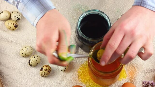 A child of Caucasian nationality paints small clouds of quail eggs with a metal spoon and lowers them into a glass jar with dyes on the Easter table. Concept for spring religious holidays