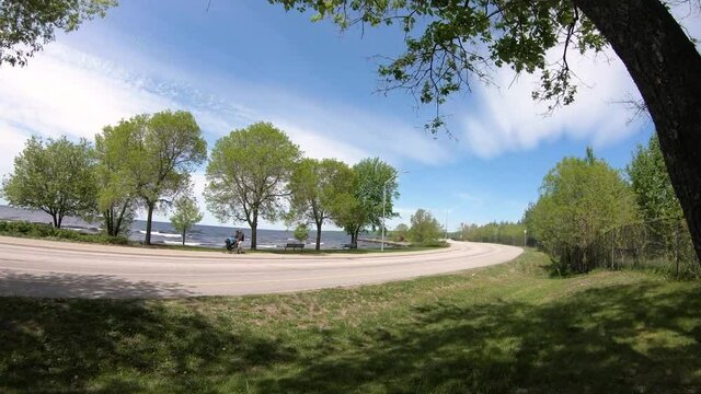 Timelapse at a waterfont on a blye sky day with many cyclists and cars going by really fast. This is a popular summer spot to go biking(Kate pace way) or to the beach in North Bay, Ontario, Canada. 