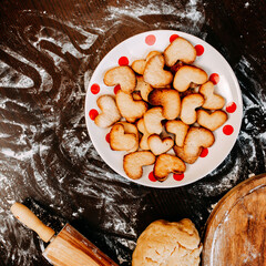 Cookies in shape of heart for the Saint Valentine's Day. Dough, flour, baking pan, round wooden cutting board and rolling pin on the table. Cookies on the plate.
