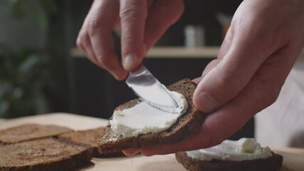 Close up shot of male hands spreading cream cheese on toasted rye bread slice with knife while cooking sandwich