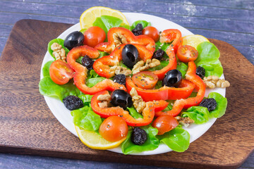 On a white plate, fresh vegetable salad with cherry tomatoes, red peppers, olives, walnuts, raisins, green salad leaves.