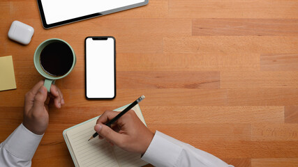 Male hand holding pencil and coffee cup while online studying with tablet and smartphone on table