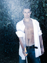Handsome male with muscular body and open shirt showering with garden hose on sunny day