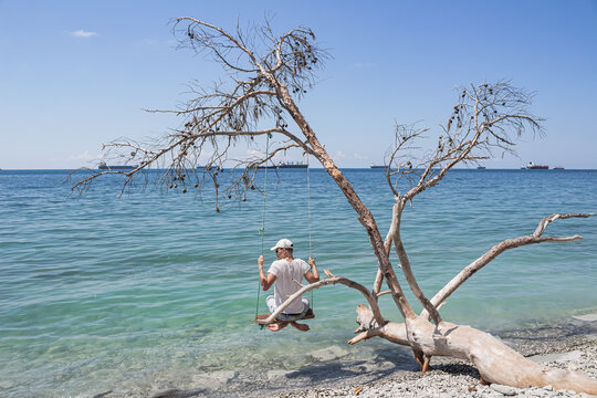 Summer landscape with a view of the sea. A guy sitting on a swing, on a fallen tree on the shore, and cargo ships on the horizon. The outskirts of the resort of Gelendzhik. Russia, Black Sea coast