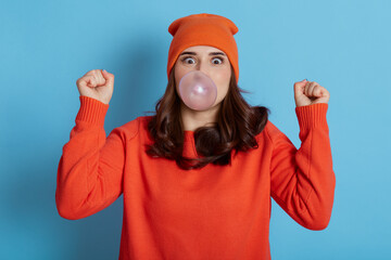 Excited dark haired female blows bubble from chewing gum, has amazed expression, dresses casual...