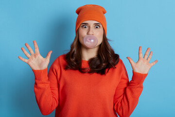 Young woman with pleasant appearance chewing gum and blowing hair bubble isolated over blue background, looks scared, being in shock, has surprise face, having fear expression, raises palms up.