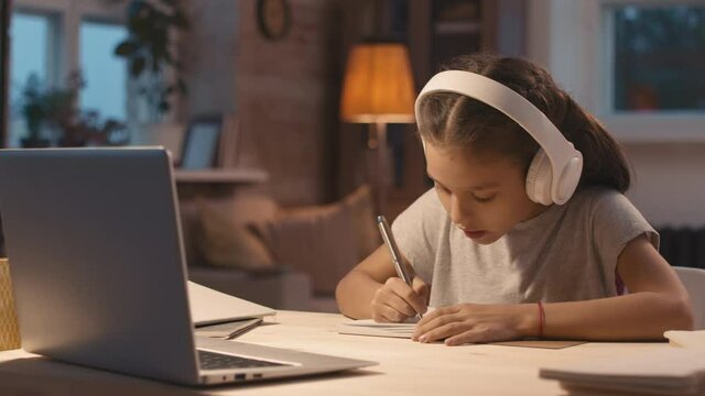 Medium shot of little girl in headphones sitting at desk at home and talking on video call with teacher while studying online