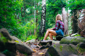 Hipster girl in the mountains. Stylish woman in checkered shirt sitting on stones in forest. Wanderlust concept. Hiking and travelling in summer. mysterious wood. Beautiful nature.