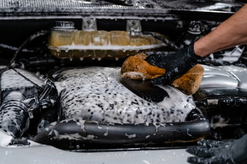 Close up of car wash worker wearing protective gloves and washing car engine with soapy sponge. Cleaning services.