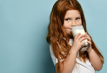 Portrait of happy red haired kid girl drinking milk or yogurt and looking at camera over blue background with free copy space. Health and diet concept. Healthy drink breakfast and clean food concept