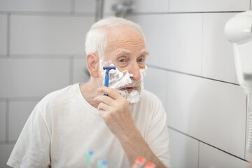 Gray-haired man in white tshirt shaving in the bathroom and looking concentrated