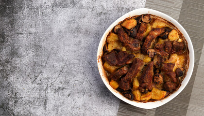 Roasted pork neck with mushrooms and potatoes in a white baking dish on a dark grey background. Top view, flat lay