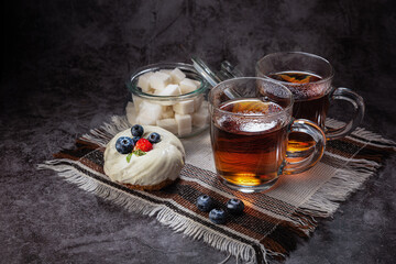 Tea in a transparent mug, with highlights, steam, with a sugar bowl with lump sugar, and a cake with strawberries and blueberries on a napkin, on a gray background