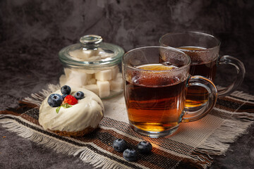 Tea in a transparent mug, with highlights, steam, with a sugar bowl with lump sugar, and a cake with strawberries and blueberries on a napkin, on a gray background