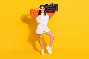 Photo portrait full body view of woman showing v-sign near eye with boombox on shoulder isolated on vivid yellow colored background