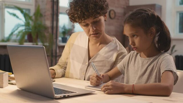 Tilt up shot of young mother with short curly hair sitting at desk beside her frustrated little daughter and explaining something to her while helping her with homework