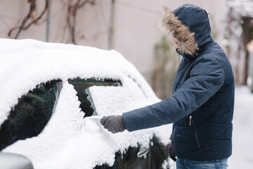 Young man cleaning snow from car windshield outdoors near the garage on winter day