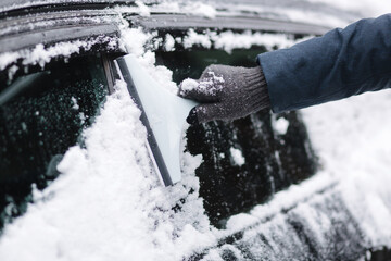 Close up of man is cleaning snowy window on a car with snow scraper. Focus on the scraper. Cold snowy and frosty morning. Black car