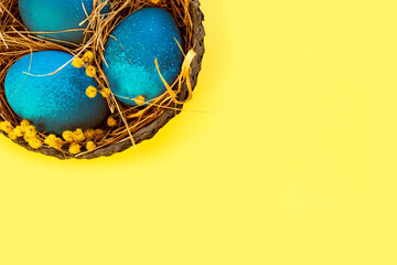 Easter egg painted bright blue lies in a nest of dry grass and flowers. Painted easter egg on a yellow background. Flat lay, top view, copy space.