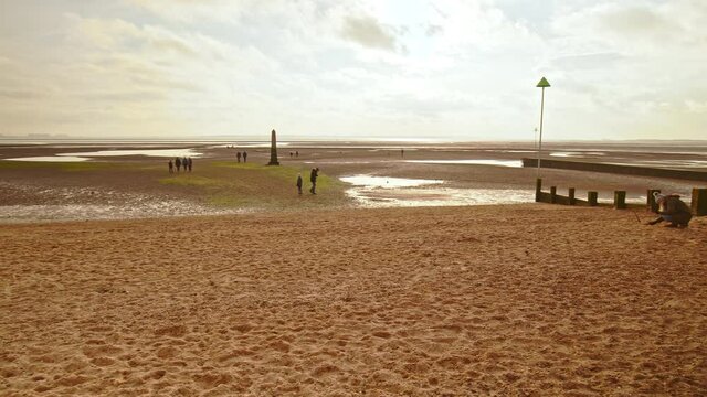 People relaxing, walking dogs on a dry beach during winter with a golden sunset. Concept of people taking care of mental health having a good time outdoors in Leigh Bell Wharf Beach, Essex, UK.