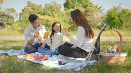 Happy family asian and little girl are drinking orange juice and have enjoyed ourselves together during picnicking on a picnic cloth in the green garden. Family enjoying sunny fall day in nature.