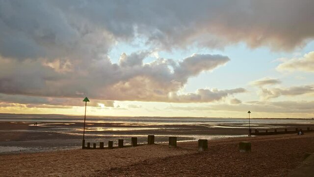 People relaxing, walking dogs on a dry beach during winter with a golden sunset. Concept of people taking care of mental health having a good time outdoors in Leigh Bell Wharf Beach, Essex, UK.