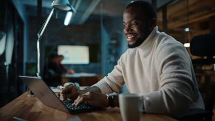 Handsome Black African American Man Having an Online Conversation on a Laptop Computer in Creative Office Environment. Happy Male is Browsing Social Media and Replying to Friends in Messenger.