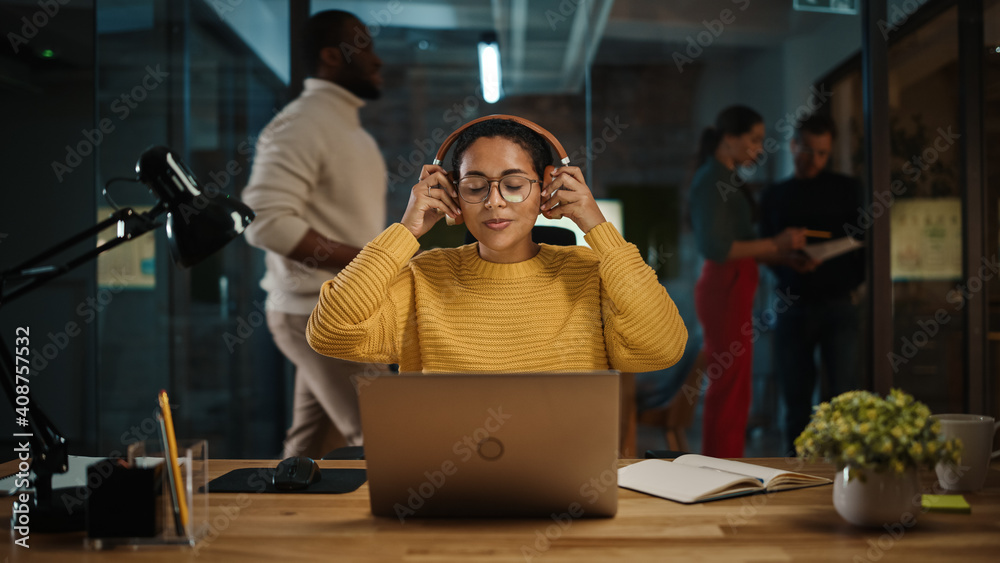 Poster young latin project manager putting on headphones and working on laptop computer in busy creative of