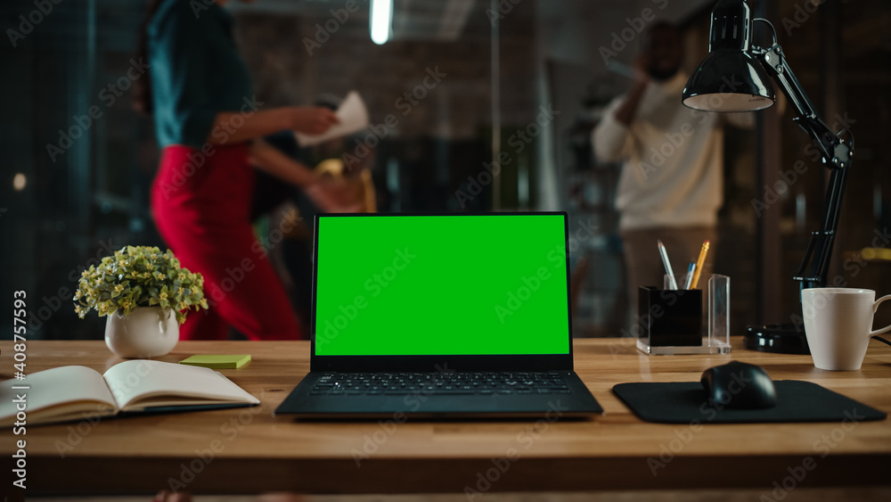 Poster Shot of a Laptop Computer with Green Screen Mock Up Display on a Desk in a Busy Creative Office Environment. Isolated Template for Any Media Type. Authentic Hipster Agency Vibe. 