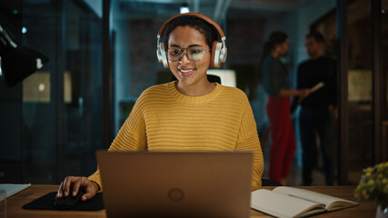 Young Hispanic Project Manager in Headphones and Working on Laptop Computer in Busy Creative Office...