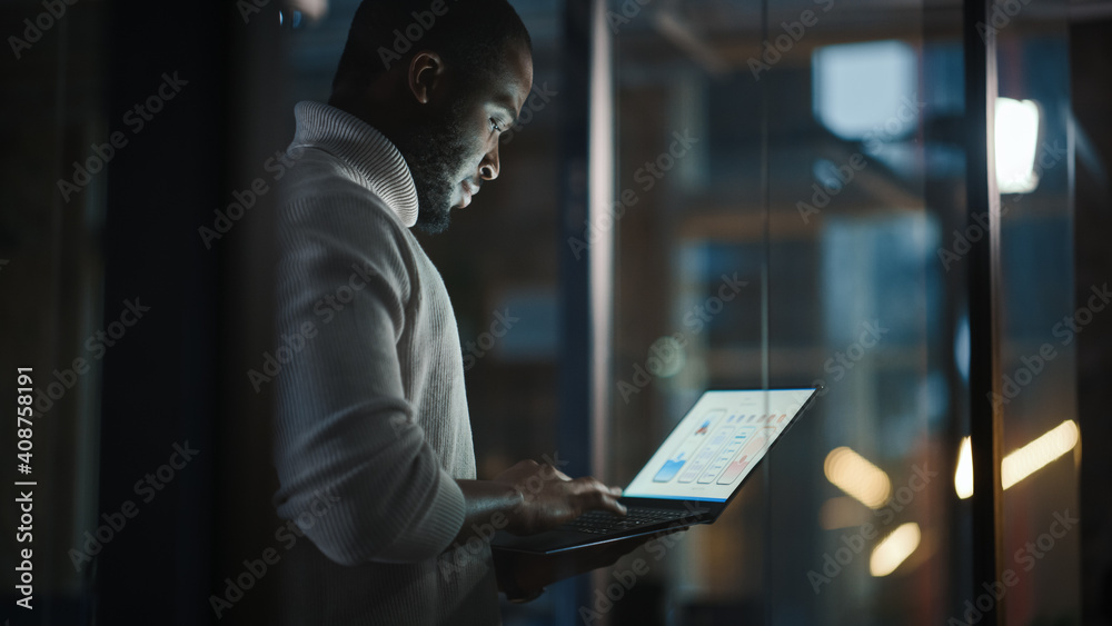 Wall mural Handsome Black African American Male is Standing in Meeting Room Behind Glass Walls with Laptop Computer in an Creative Agency. Project Manager Wearing White Jumper and Working on App User Interface.