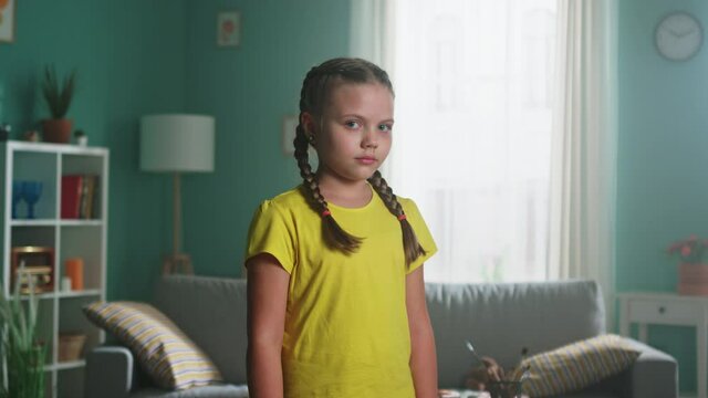 Portrait of small sad girl with two braids and green eyes, in yellow T-shirt, standing at home and looking at camera, feeling unhappy and unloved, Foreground, Slow motion.