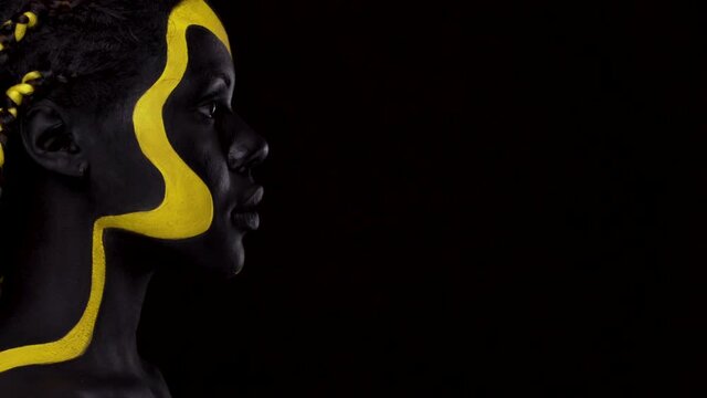 Face art. Woman with black and yellow body paint. Young african girl with colorful bodypaint. An amazing afro american model with yellow makeup. Closeup face.